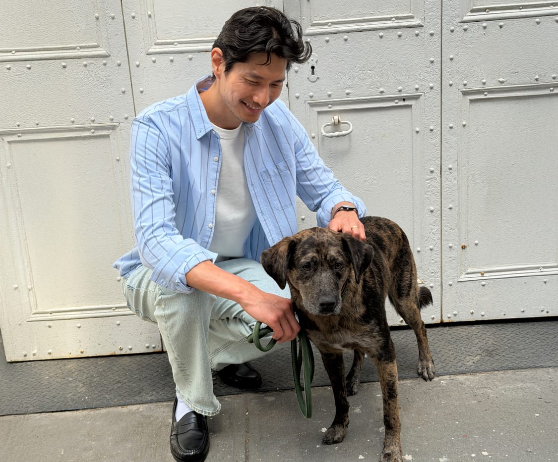 Man sitting while petting a dog, wearing a light blue vertical-striped button-down shirt over a white shirt and jeans