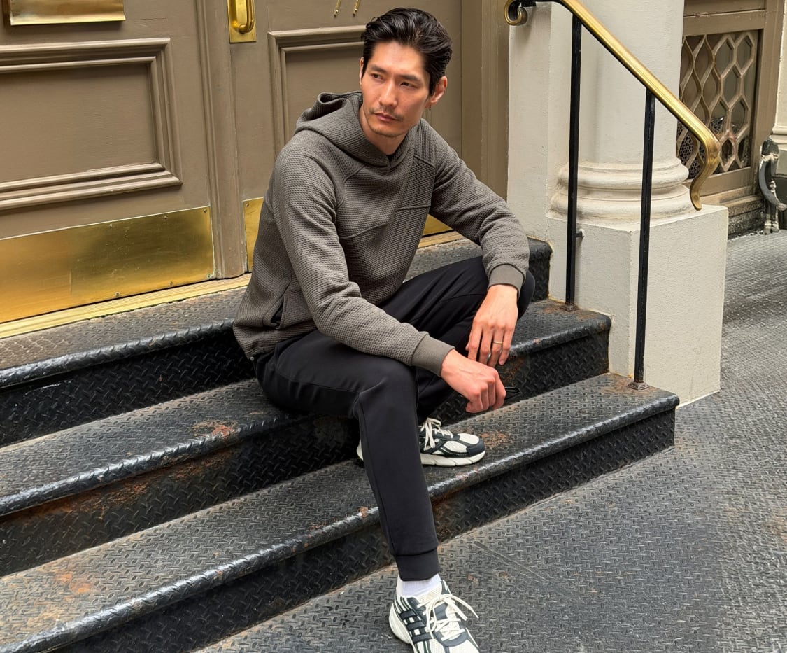 Man sitting on outdoor stairs wearing a grey textured pullover hoodie, dark pants, and grey and white athletic sneakers