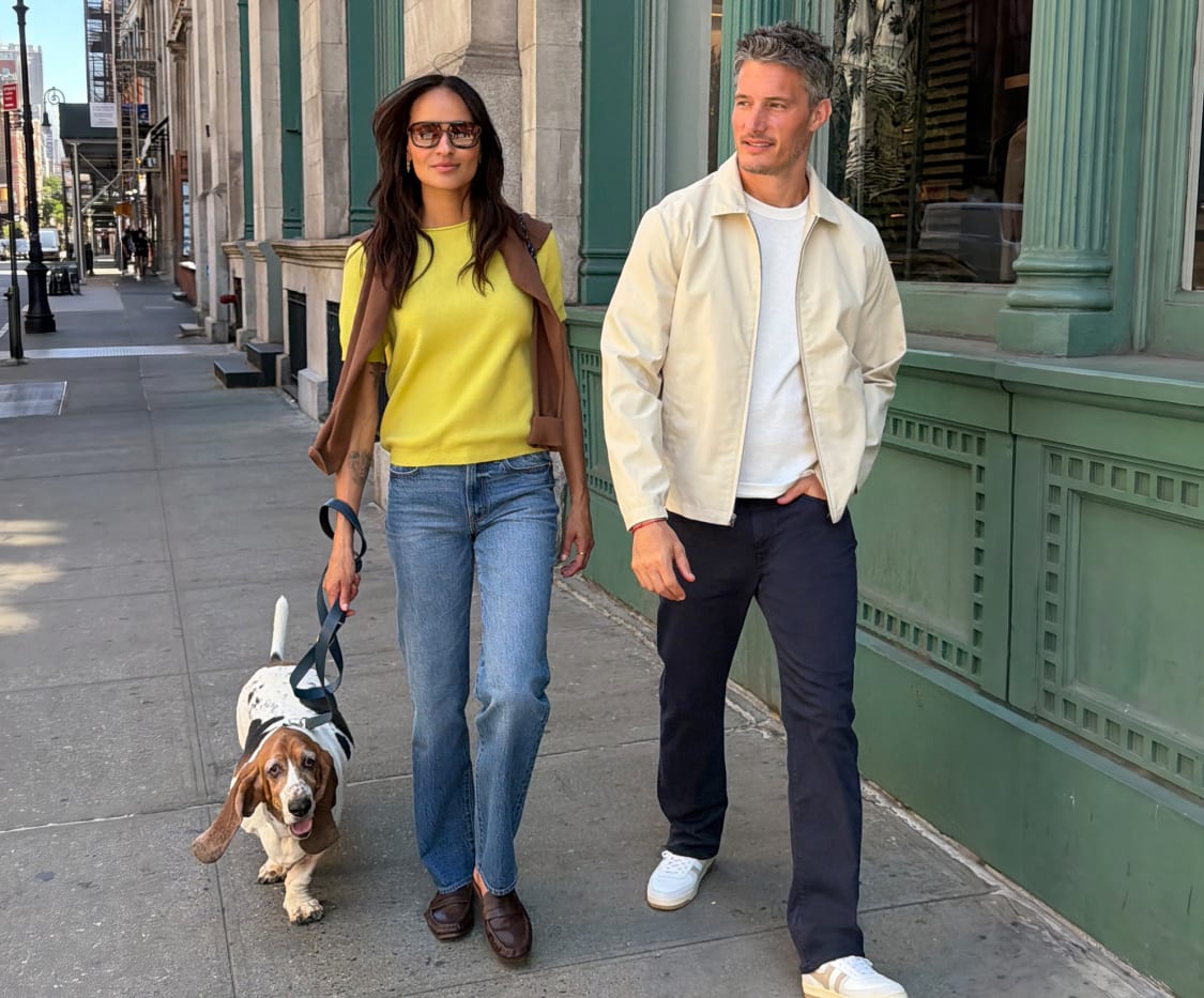 Woman in yellow top and jeans alongside a man in cream zip jacket and dark trousers walking a dog on a sidewalk