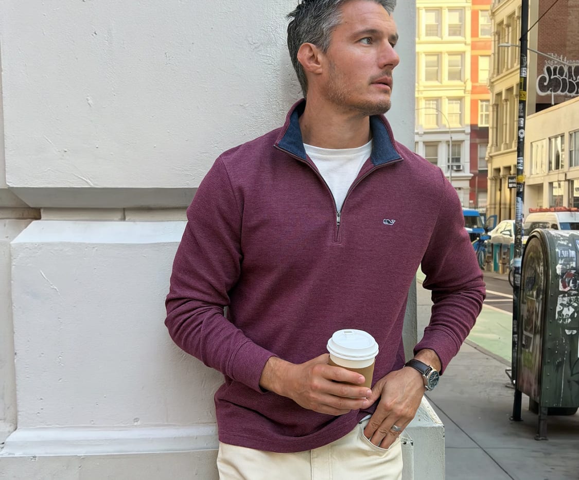 Man wearing a dark plum quarter-zip pullover sweater over a white t-shirt and cream-colored pants while holding a coffee cup