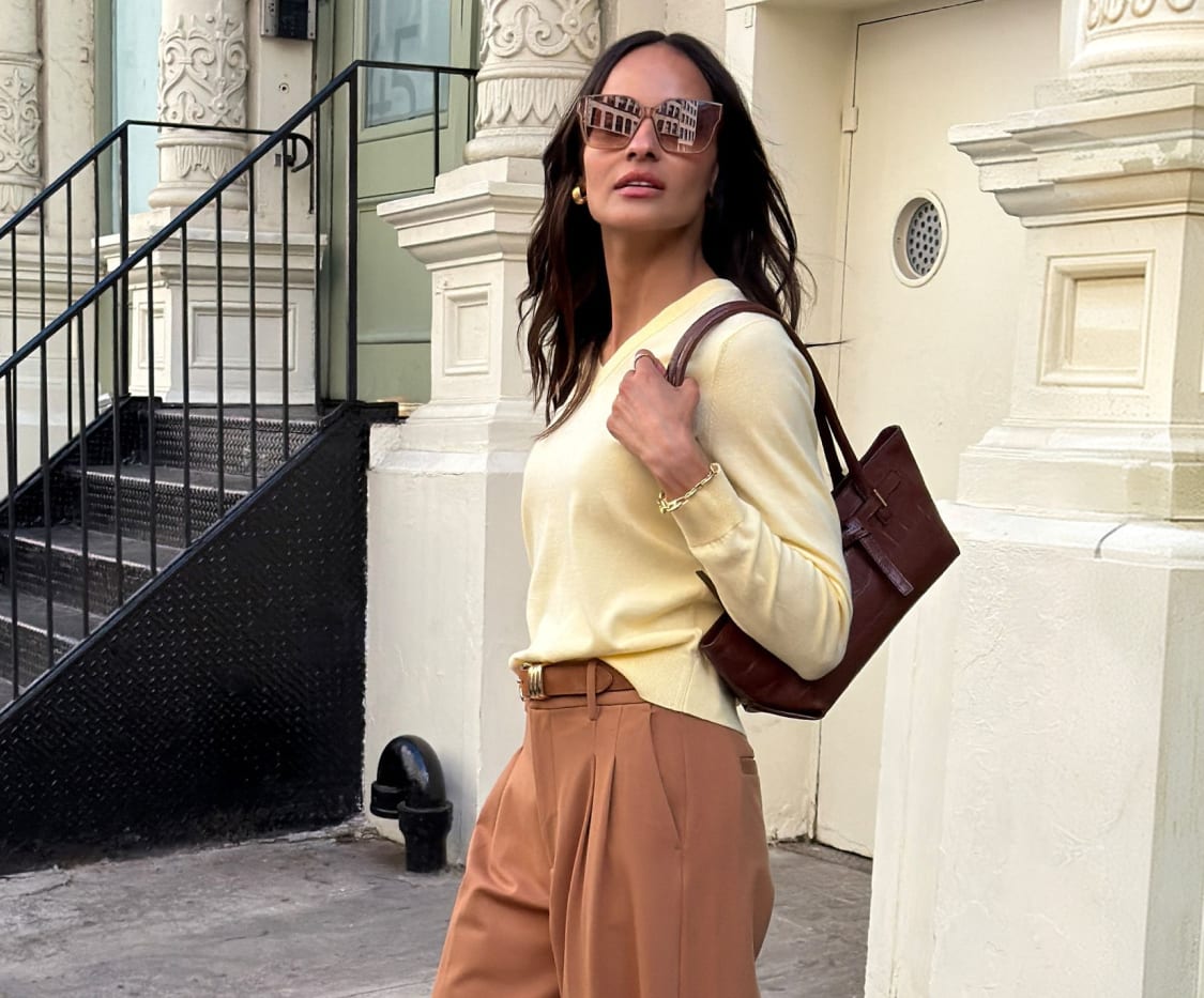 Woman wearing a pale yellow sweater, light brown wide-leg trousers, sunglasses, carrying a dark brown handbag