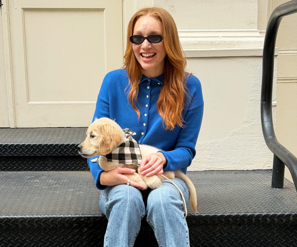 Woman sitting in a royal blue polo cardigan sweater, light blue jeans, black loafers, holding a small dog