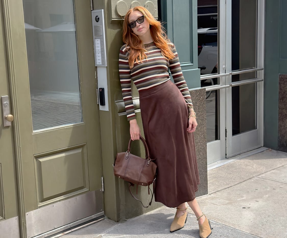 Woman in a long-sleeve olive, brown, and cream striped sweater, long brown skirt, sunglasses, carrying a brown bag