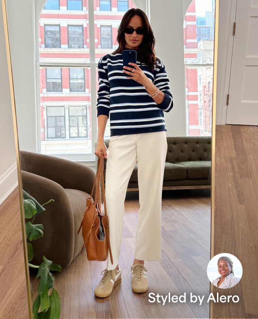 Woman wearing a blue pinstripe button-up shirt, grey sweater, dark blue jeans, tan heels, and black tote bag