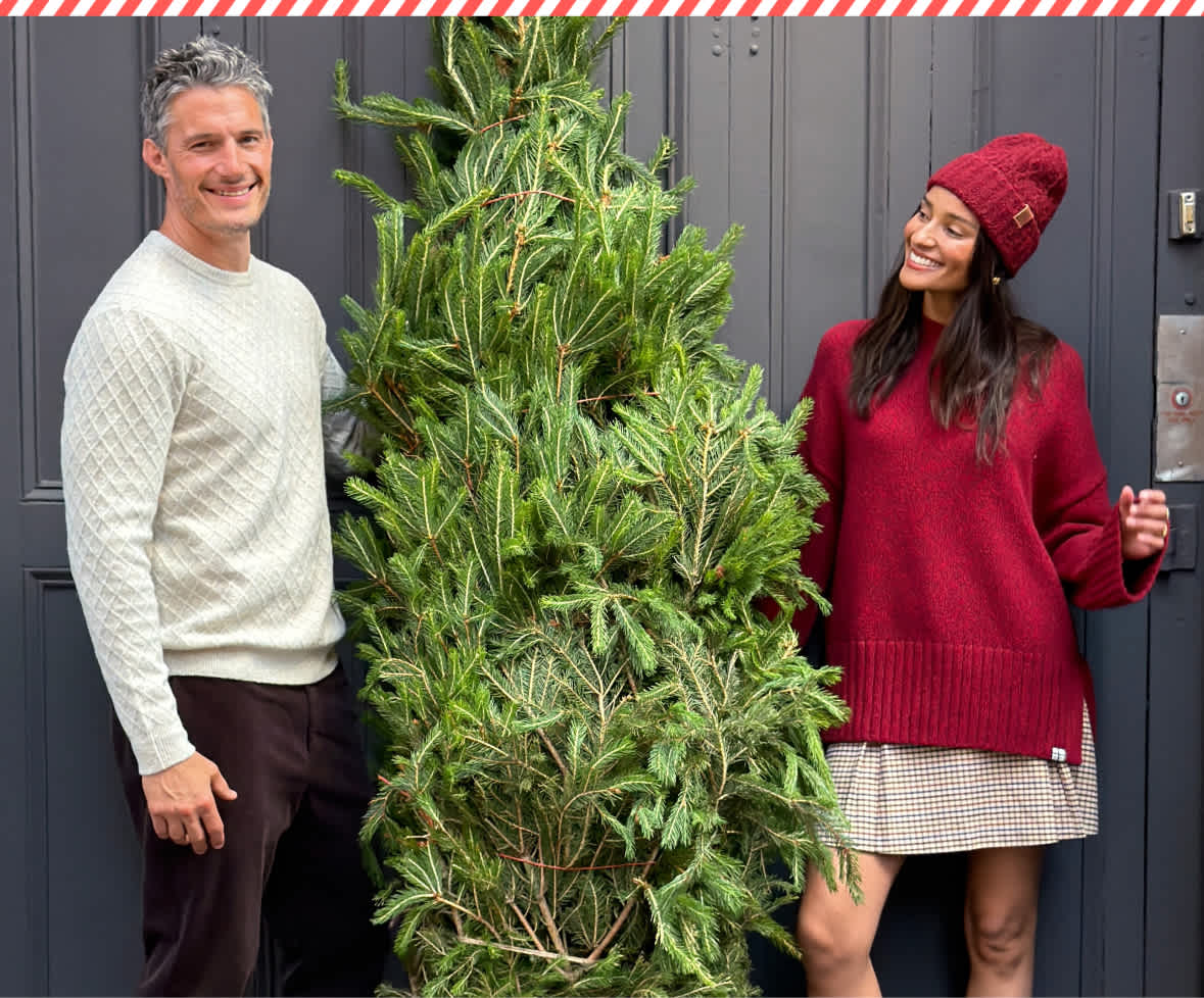 A split holiday image showing a man in a white sweater and a woman in a red sweater standing on a city sidewalk next to a Christmas tree on the left, and a smiling woman wearing a yellow beanie and camel coat holding stacked Stitch Fix gift boxes tied with teal ribbons on the right.