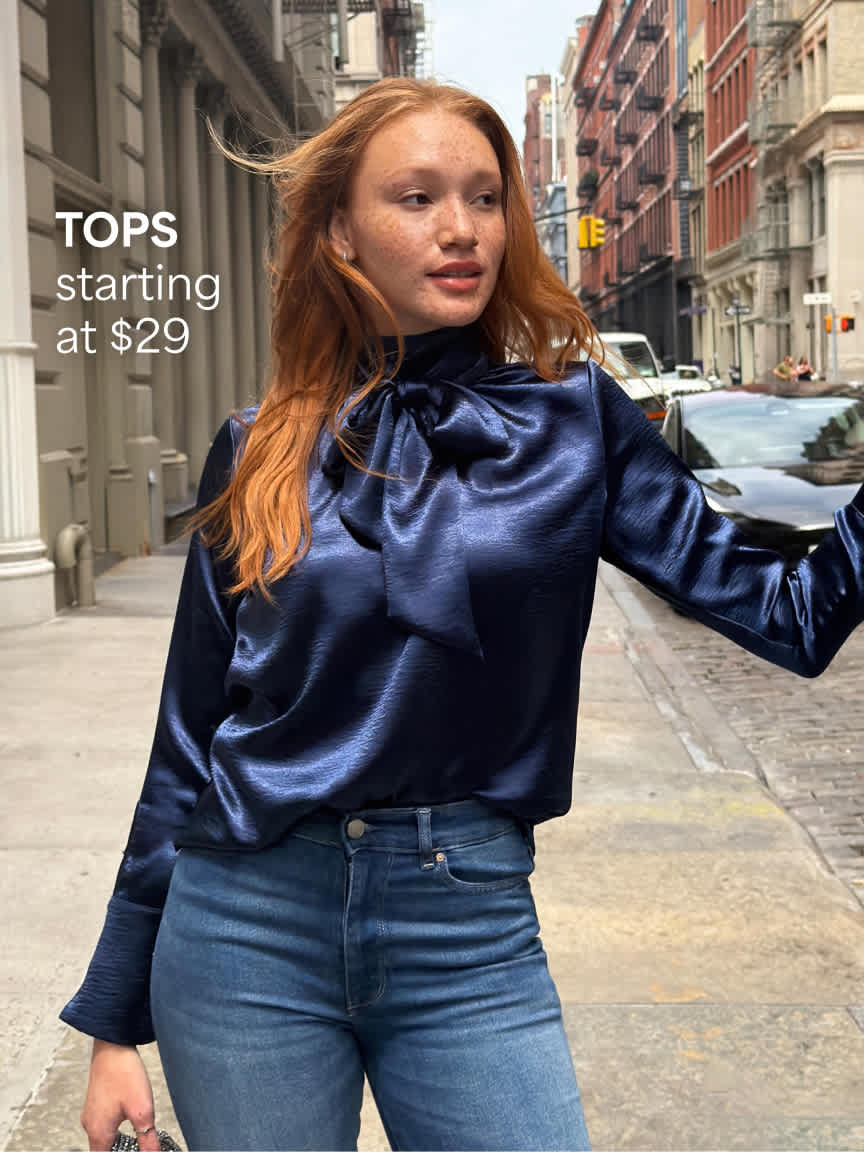 A redheaded woman walking down a city street wearing a lustrous navy blue satin high-neck tie blouse paired with high-waisted blue jeans. Text overlay reads "TOPS starting at $29".