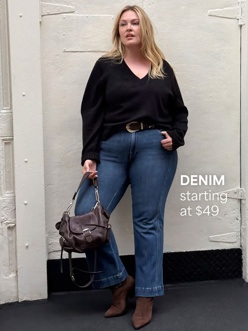 A blonde woman standing against a white building wall wearing a long-sleeve black v-neck sweater, classic blue bootcut jeans with a dark belt, dark brown ankle boots, and holding a brown shoulder bag. Text overlay reads "DENIM starting at $49".