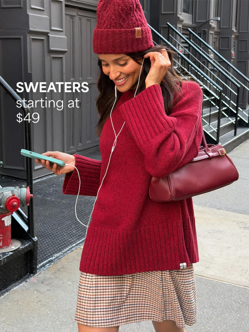 A smiling woman walking outdoors holding a phone and wearing a dark red beanie, an oversized deep red knit sweater over a plaid skirt, and carrying a matching red leather handbag. Text overlay reads "SWEATERS starting at $49".