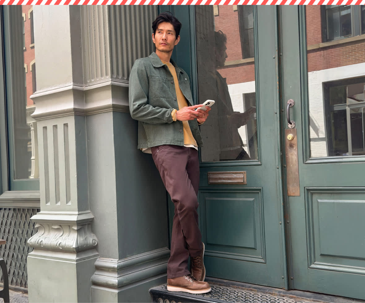 A split lifestyle image featuring a man leaning against a green doorway holding a smartphone while wearing an olive green jacket and burgundy trousers on the left, and a man sitting on outdoor steps holding a coffee cup, wearing a burgundy quarter-zip pullover and cream khakis on the right.