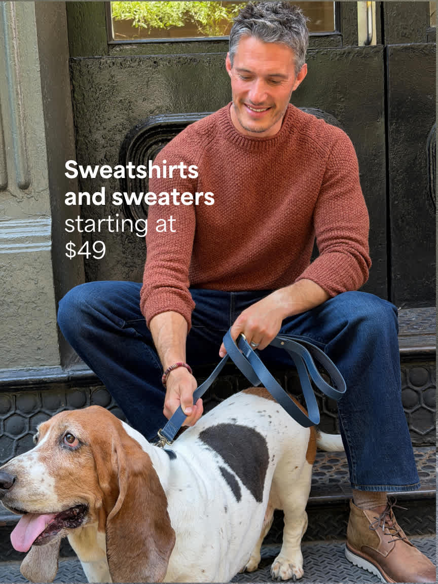 A man with grey hair sitting on outdoor stone steps holding a leash attached to a basset hound, wearing a rust-colored knit sweater, dark blue jeans, and brown leather boots. Text overlay reads "Sweatshirts and sweaters starting at $49".