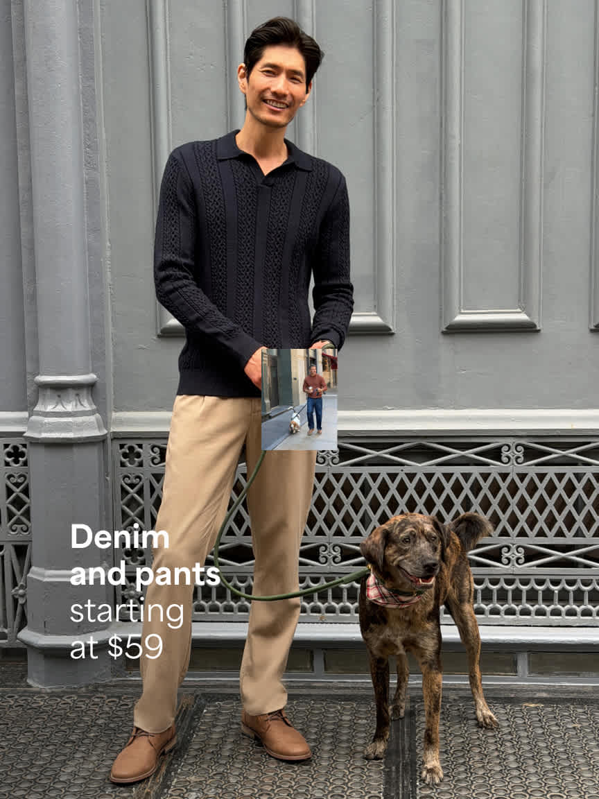 A man standing outdoors on a city sidewalk holding a dog leash attached to a brindle dog, wearing a dark navy long-sleeve polo knit, beige trousers, and tan shoes. Text overlay reads "Denim and pants starting at $59".