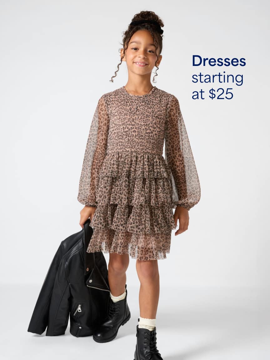 A young girl with an updo hairstyle standing against a white background wearing a brown tiered chiffon long-sleeve dress and black boots, holding a black leather jacket. Text overlay reads "Dresses starting at $25".