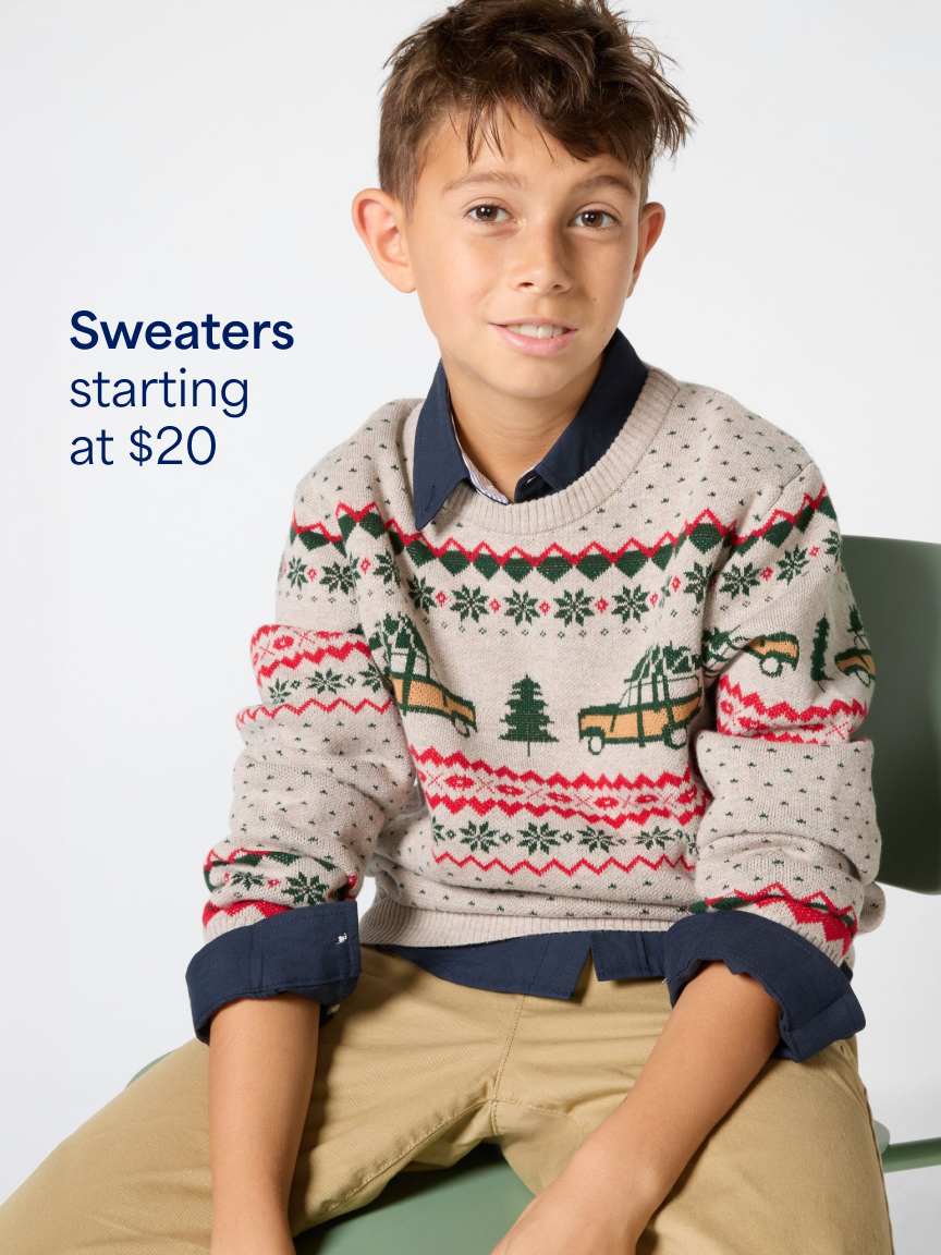 A young boy sitting on a chair wearing a festive fair-isle knit sweater with trees and trucks layered over a dark collared shirt and tan pants. Text overlay reads "Sweaters starting at $20".