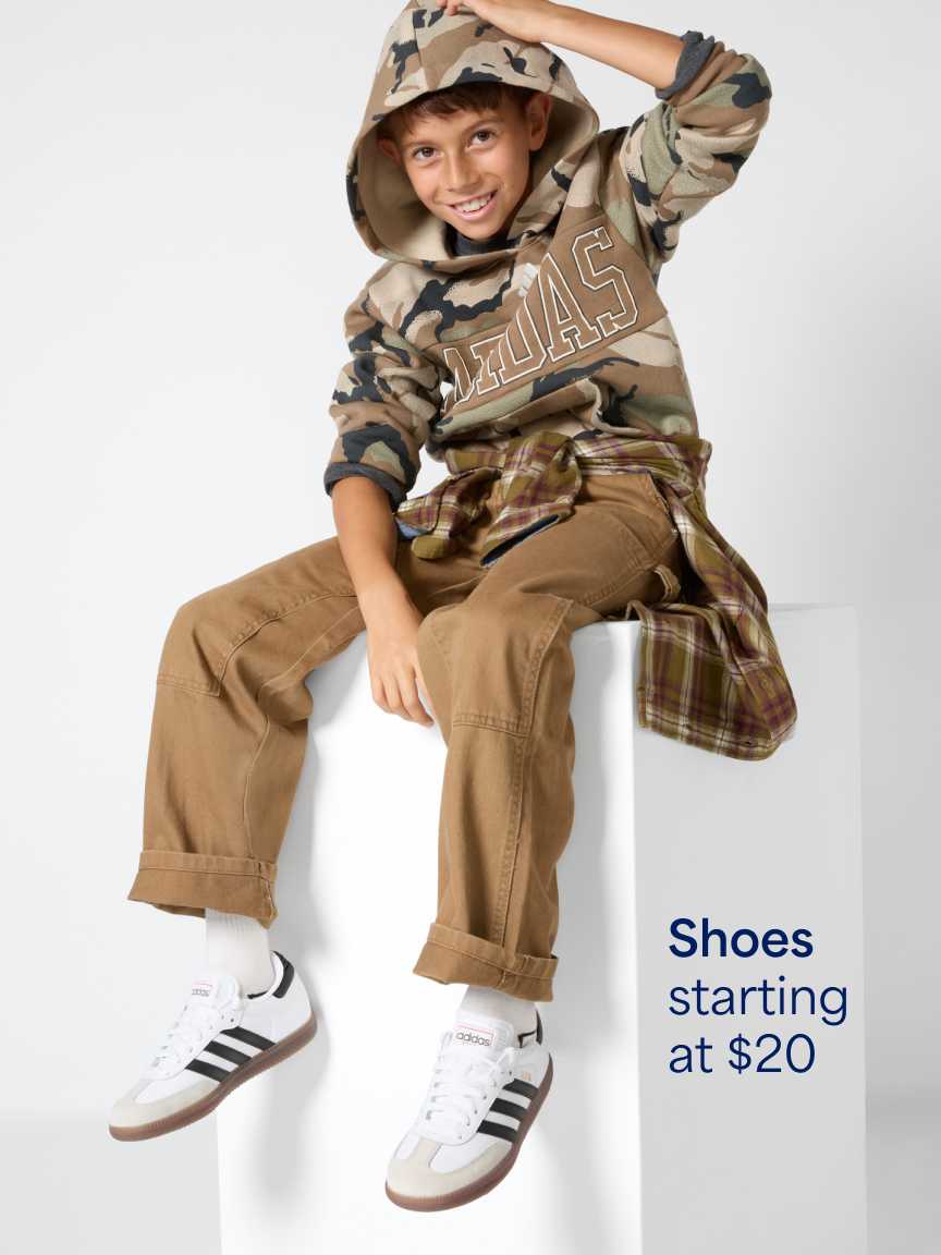A young boy sitting on a white block wearing a camo hooded jacket over a long plaid shirt, tan cargo pants, and white Adidas sneakers with black stripes. Text overlay reads "Shoes starting at $20".