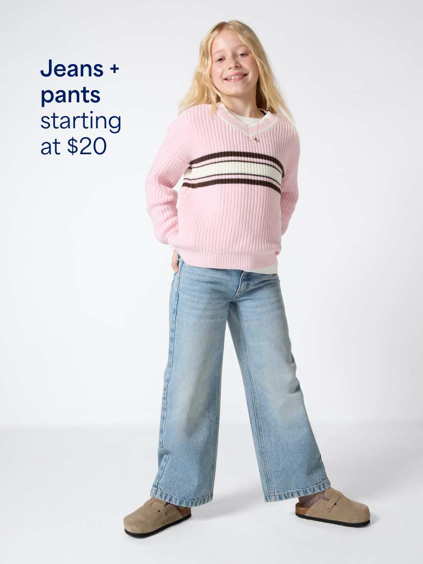 A young blonde girl posing in a light pink sweater with a striped chest band, light-wash wide-leg denim jeans, and tan suede clogs. Text overlay reads "Jeans + pants starting at $20".