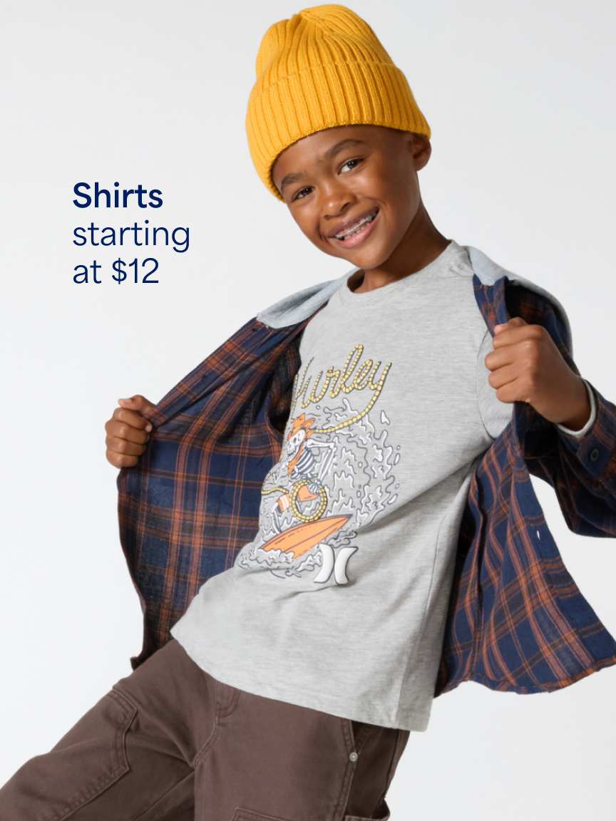 A smiling young boy wearing a yellow beanie, a grey graphic t-shirt open over a plaid unbuttoned shirt, and brown pants. Text overlay reads "Shirts starting at $12".