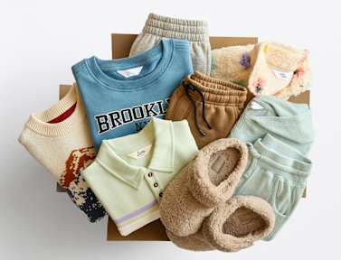 Stitch Fix box opened with folded kids’ clothes coming out of it. Featured items include: a blue graphic sweatshirt, gray jogger pants, sherpa jacket, a mint matching lounge set, a graphic sweater and fuzzy tan slippers.