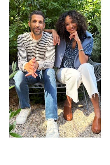 Stitch Fix models, a man and woman, sitting next to each other on a bench wearing fresh outfits. The man is in a white and black striped sweater, blue jeans and retro-looking sneakers. The woman is wearing a blue floral blouse with a blue sweater over her shoulders, white cut-off jeans and tan clogs.