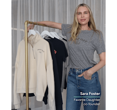 Woman wearing a heather gray "Drop Off Club" graphic T-shirt and blue jeans stands beside a clothing rack displaying coordinating Favorite Daughter apparel, including a cream quarter-zip sweatshirt and graphic T-shirts. White text overlay identifies Sara Foster, Favorite Daughter co-founder.