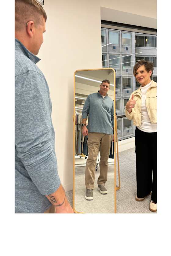 Model wearing Stitch Fix big and tall men’s clothing, including: light blue quarter-zip and khaki pants, looks at his outfit in a mirror. A woman nearby gives a thumbs-up.