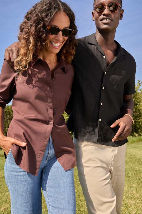 Two adults in effortless casual style walk arm in arm outdoors, showcasing versatile everyday pieces including a brown button-front shirt, light-wash jeans, a black short-sleeve shirt, cream pants, and sunglasses.