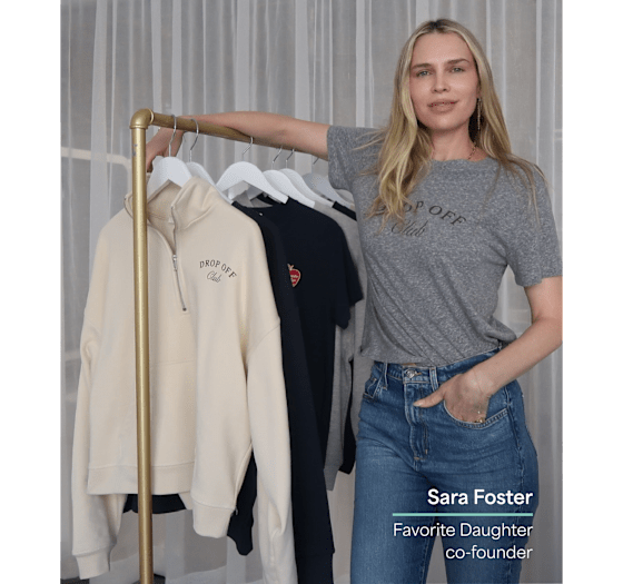 Woman wearing a heather gray "Drop Off Club" graphic T-shirt and blue jeans stands beside a clothing rack displaying coordinating Favorite Daughter apparel, including a cream quarter-zip sweatshirt and graphic T-shirts. White text overlay identifies Sara Foster, Favorite Daughter co-founder.