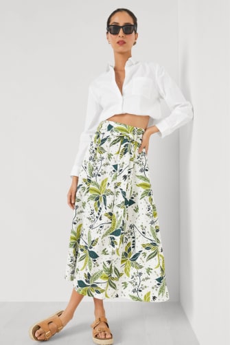 A female model wears black oversized sunglasses, a cropped white cotton shirt, and a high-waisted green and white palm print full midi skirt and tan sandals. 
