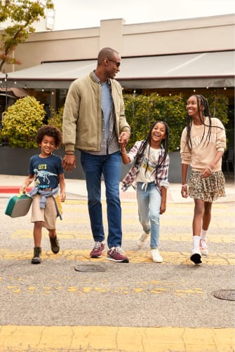 Models wearing Stitch Fix clothing including a dad in a khaki bomber jacket and jeans, three kids in casual clothes—boy in dinosaur T-shirt and shorts, younger girl in jeans and a striped sweater and older girl in a floral skirt, pink sweatshirt and sneakers.