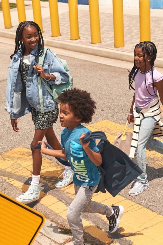 Three kids in crosswalk, one wearing floral dress, denim jacket and white sneakers from Stitch Fix, one wearing purple t-shirt, grey sweatpants and striped blue and white sweater, one wearing blue graphic t-shirt and grey sweatpants